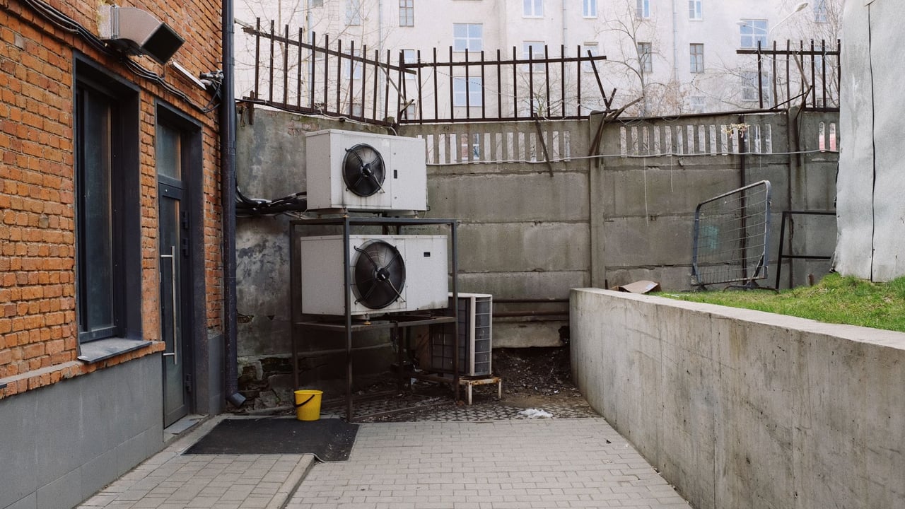 Contemporary building from brick with white air conditioning systems and yellow bucket in yard with fence and concrete wall in city