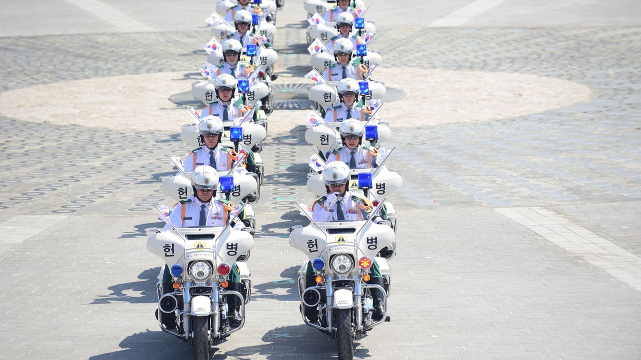 A line of South Korean police officers on motorcycles holding flags in an urban parade.