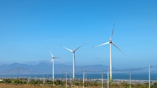 Scenery view of wind turbines in row on terrain with plants against ocean and mounts in daytime