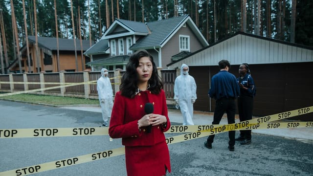 Journalist reporting from a crime scene with STOP tape and forensic team in the background.