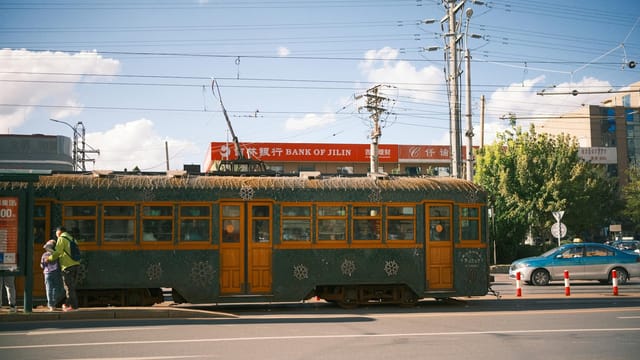 Vintage tram travels through city street near Bank of Jilin under clear skies.
