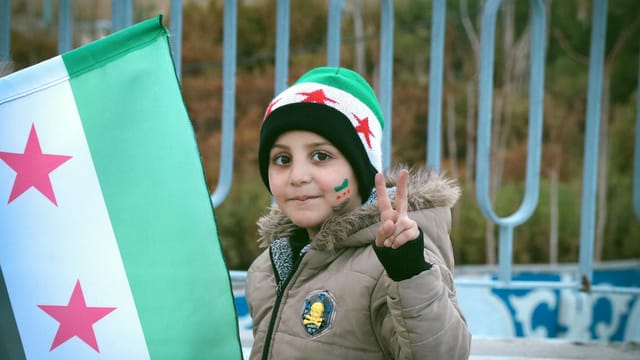 A young child in Syria showing a peace sign beside the Syrian Independence flag, symbolizing hope and freedom.