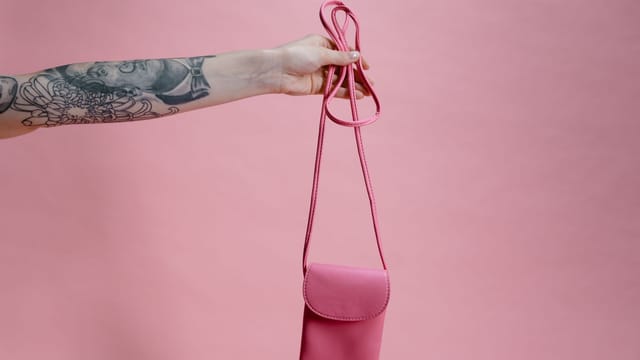 A tattooed arm elegantly holds a pink sling bag against a pink background, showcasing style and fashion.