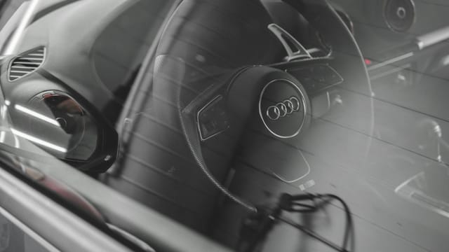 Photo of Audi car interior through the window focusing on the steering wheel and dashboard.