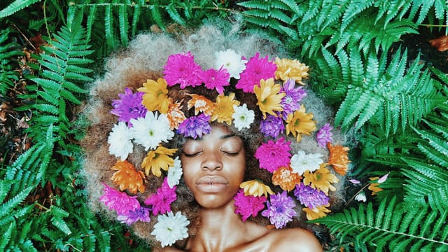A serene portrait of a woman with flowers in her afro, surrounded by lush green ferns.