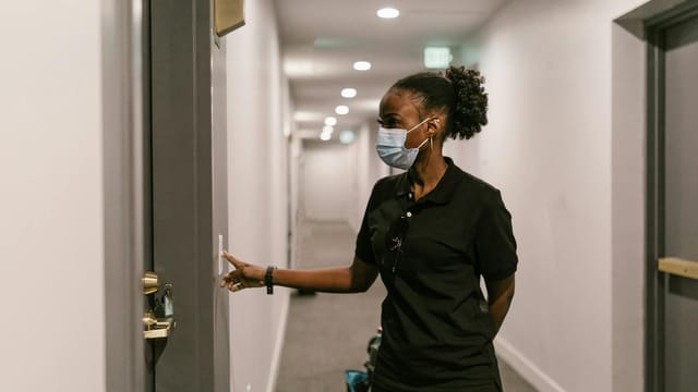 Delivery woman wearing a face mask presses doorbell in a hallway, signifying logistics and online shopping.