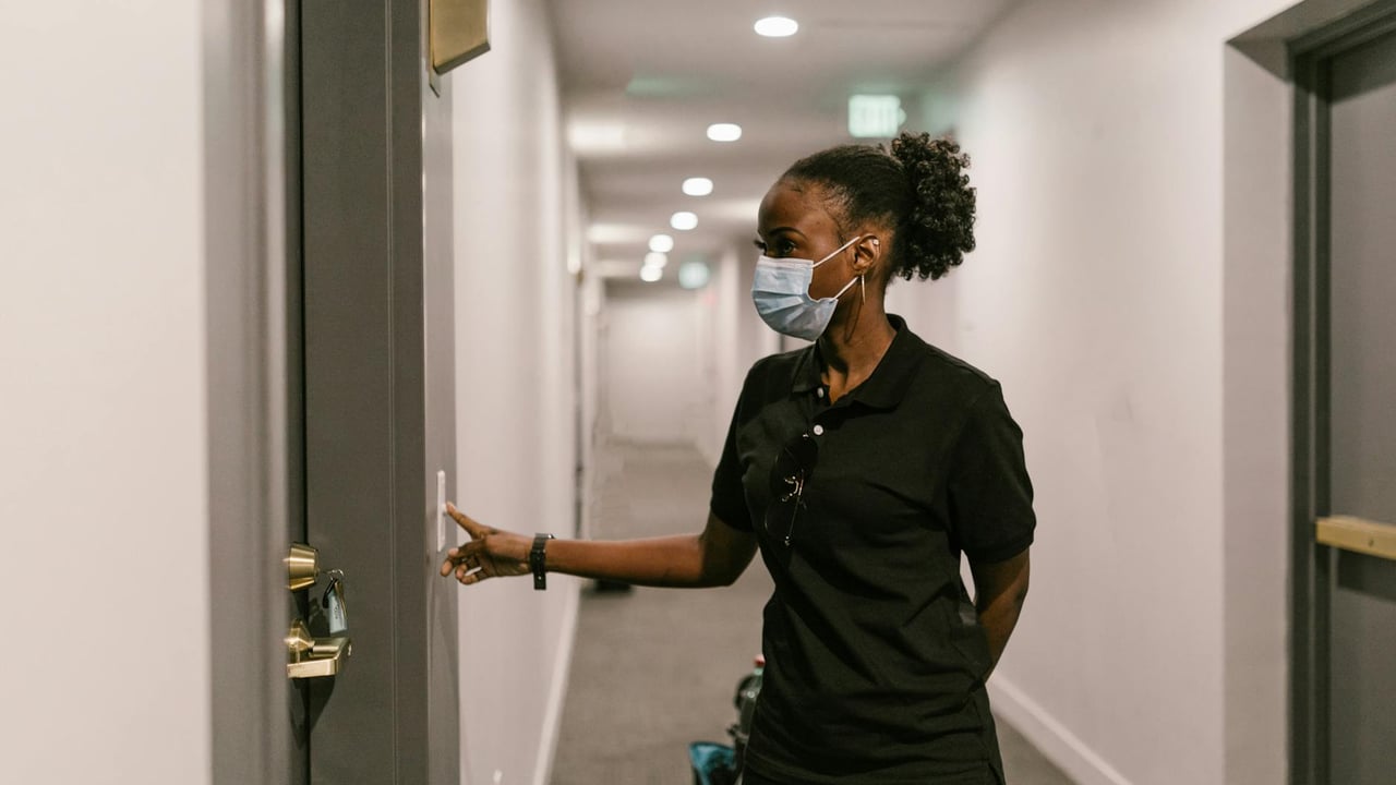 Delivery woman wearing a face mask presses doorbell in a hallway, signifying logistics and online shopping.
