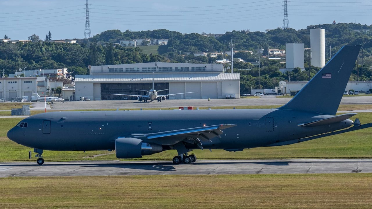 A military aircraft taxiing on a runway at an airbase surrounded by lush green hills.