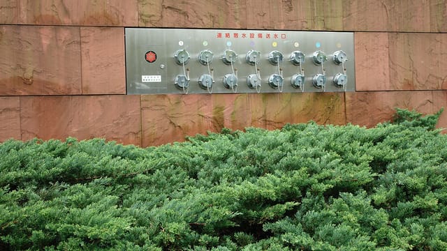 Metal valve panel integrated into a textured wall surrounded by greenery, Japan.