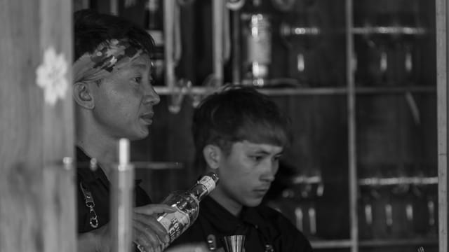 Two men with short hair and hairband preparing drinks in a black-and-white bar setting.