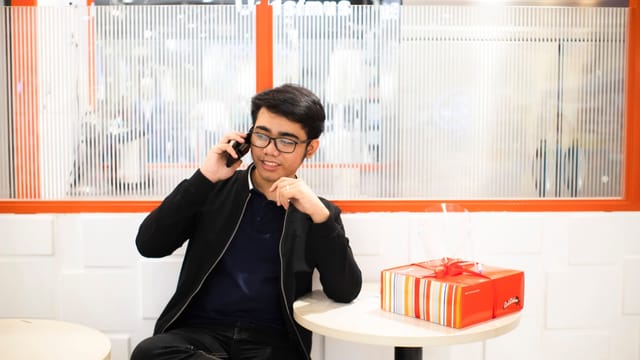 Confident young ethnic guy in casual clothes sitting near table with present box and window while having phone call in light mall