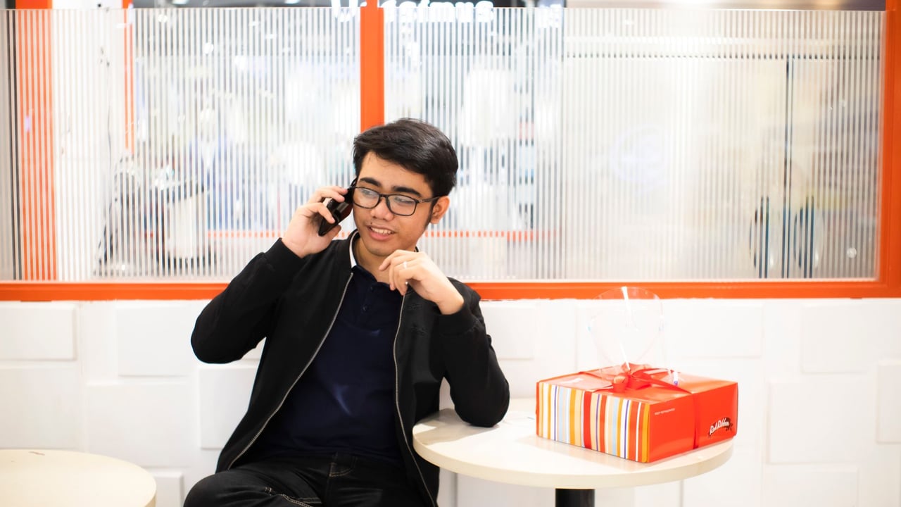 Confident young ethnic guy in casual clothes sitting near table with present box and window while having phone call in light mall