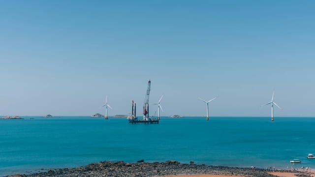 A serene view of offshore wind turbines against a clear blue sky with calm ocean waters.