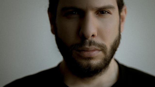 Close-up portrait of a bearded man with a serious expression set against a simple background.