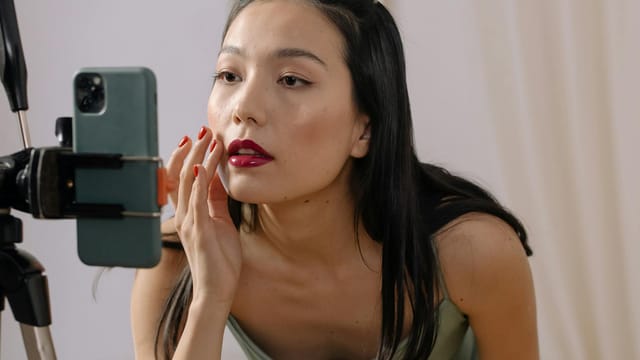 Asian woman applying red lipstick during a live streaming makeup vlog using a smartphone.