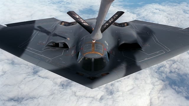 A stealth bomber aircraft receives aerial refueling mid-flight over clouds.