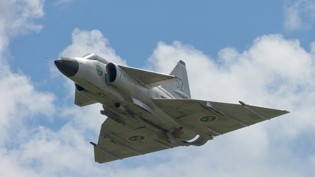 A sleek Swedish fighter jet flying through a bright blue sky with fluffy clouds.