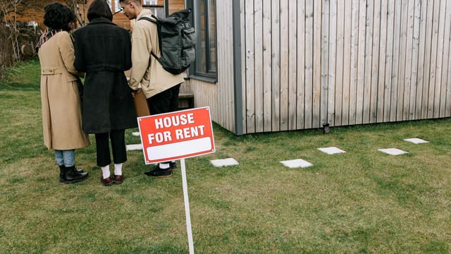 Group of adults viewing a wooden house with a 'House for Rent' sign on the lawn.